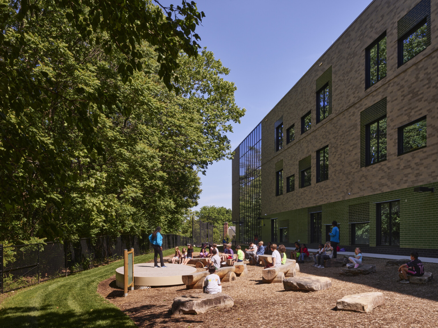 Outdoor learning area next to a multistory school filled with rocks and cut log seating looking at a densely wooded area