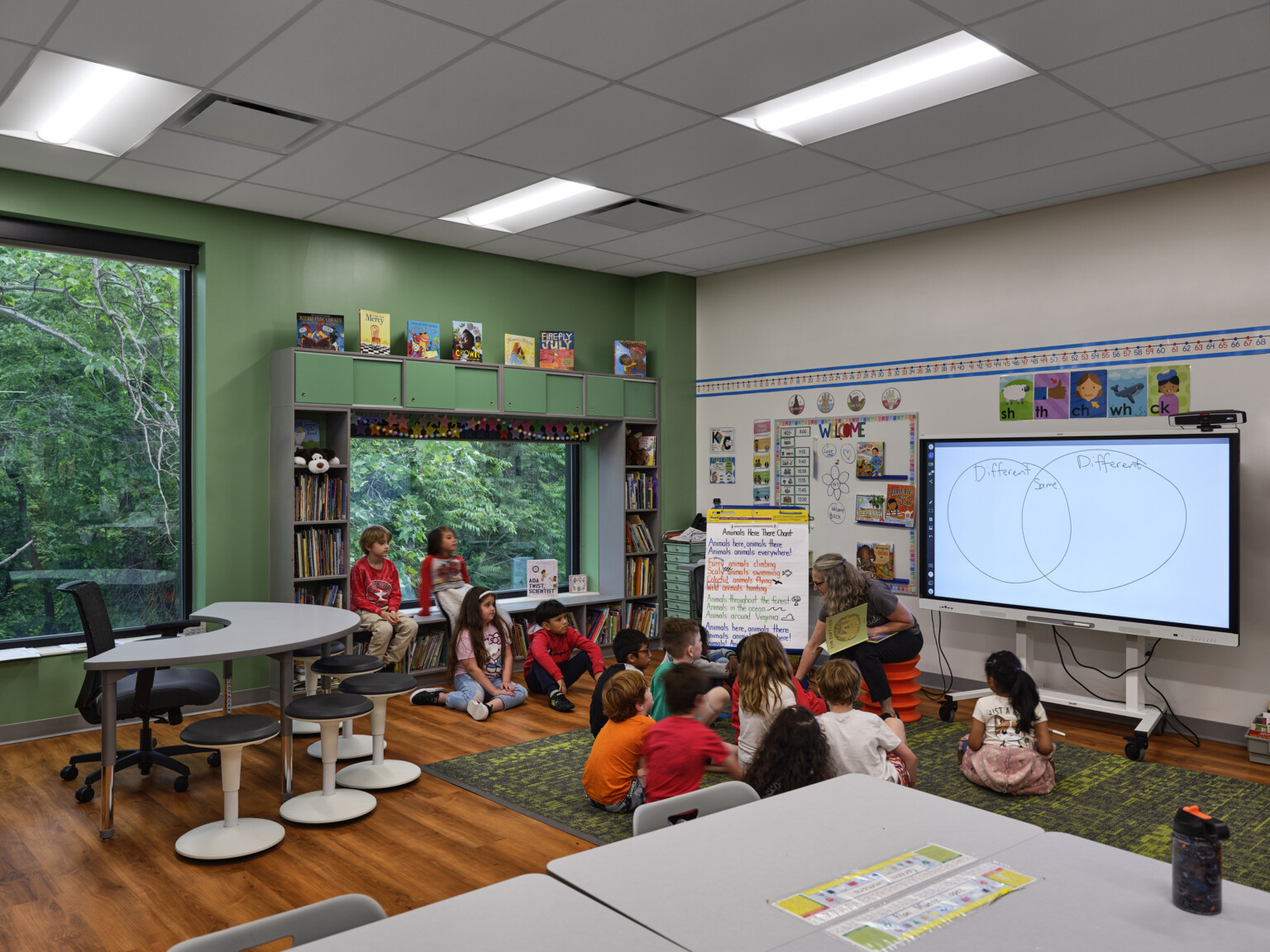 Elementary school classroom filled with students in front of a large monitor next to a green accent wall filled with large windows