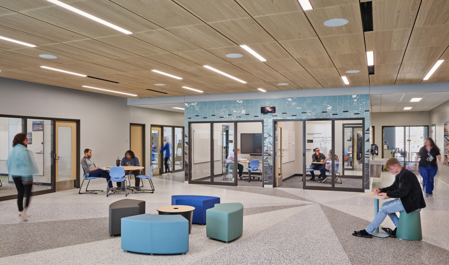 Open concept gathering area with blue and green modern seating filled with students surrounded by conference rooms with glass doors