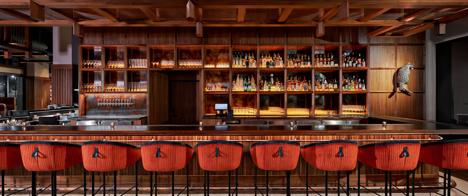 Large wood bar with built in shelves along the back. Wood overhang over back of bar, and stools with red and black upholstery