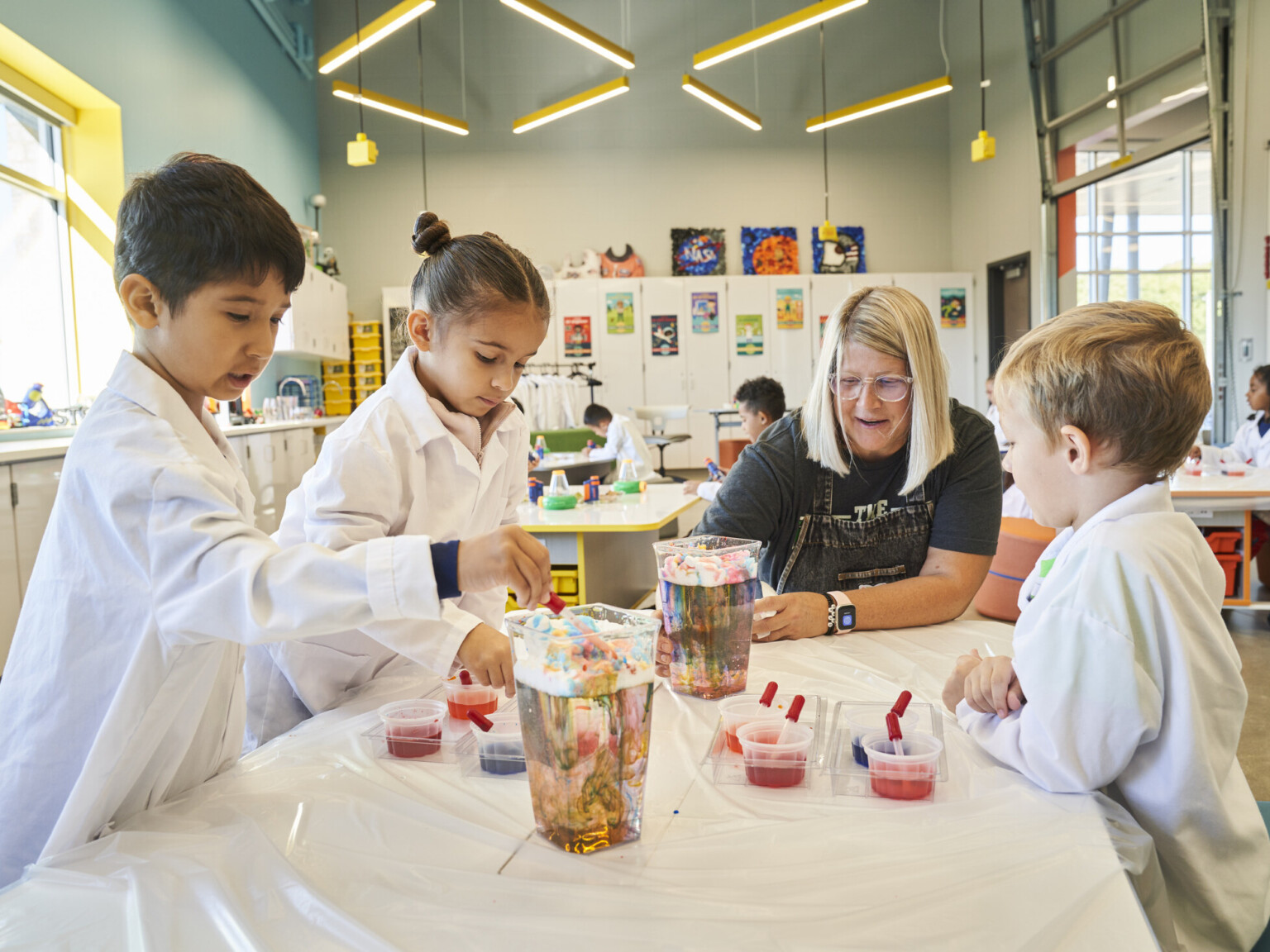 A group of kids in lab coats working with their teacher on a science experience in a lively science classroom