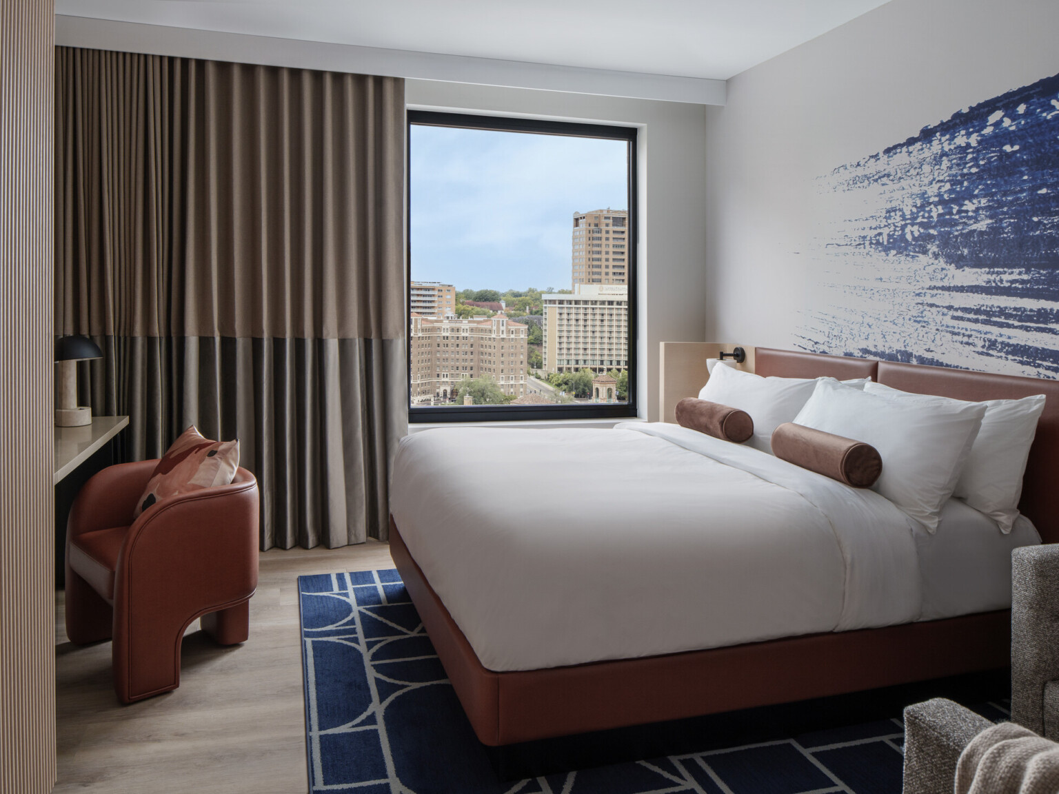 Hotel room with blue artwork behind bed with white linens and dark orange furniture