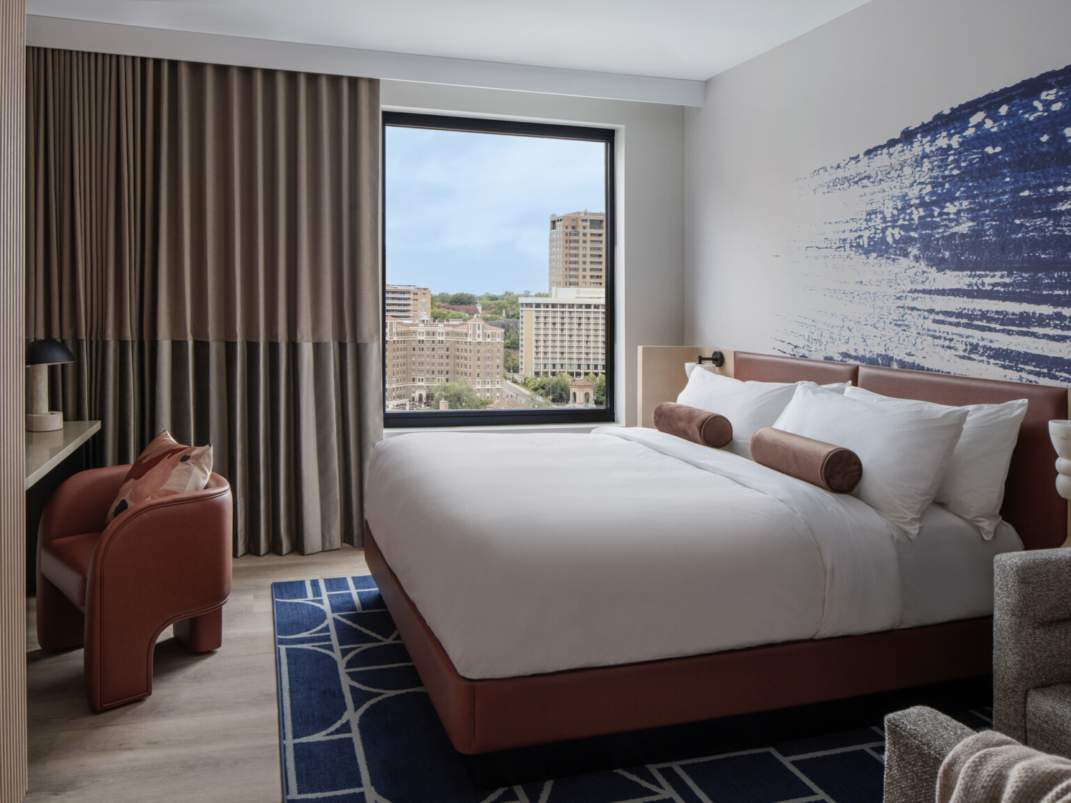 Hotel room with blue artwork behind bed with white linens and dark orange furniture