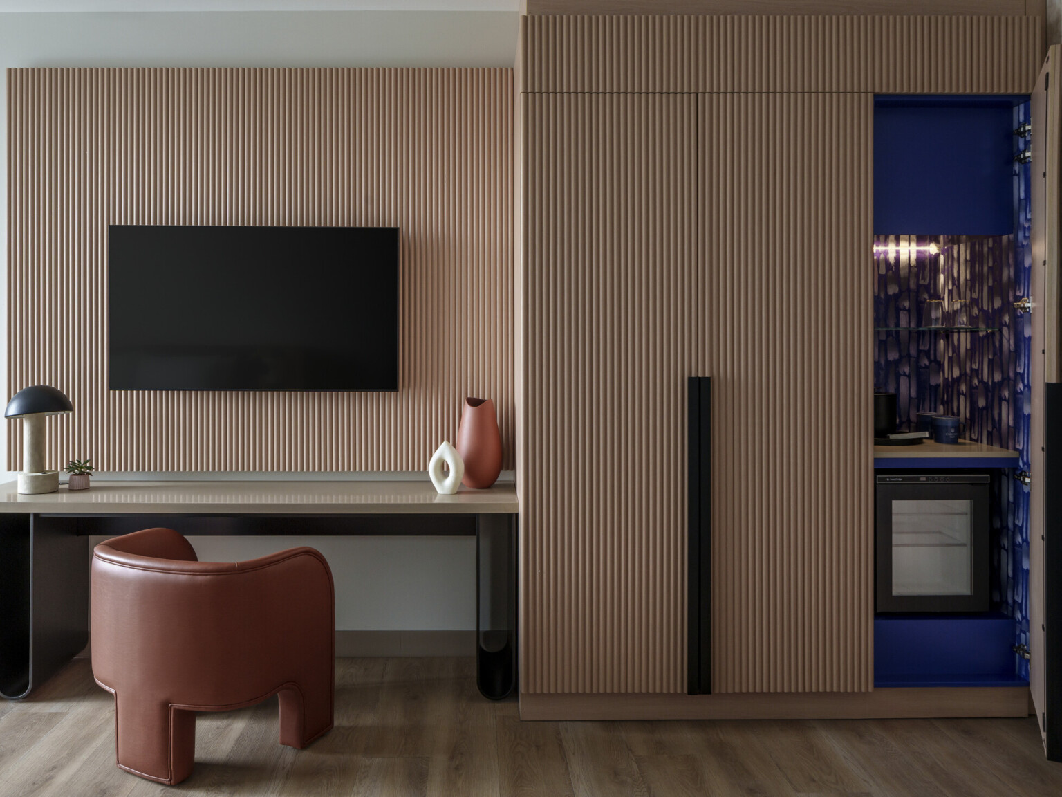 Modern hotel room desk area showing a wooden accent back drop, desk with chair and TV hanging on wall and cabinets to the right with large door open showing blue accents