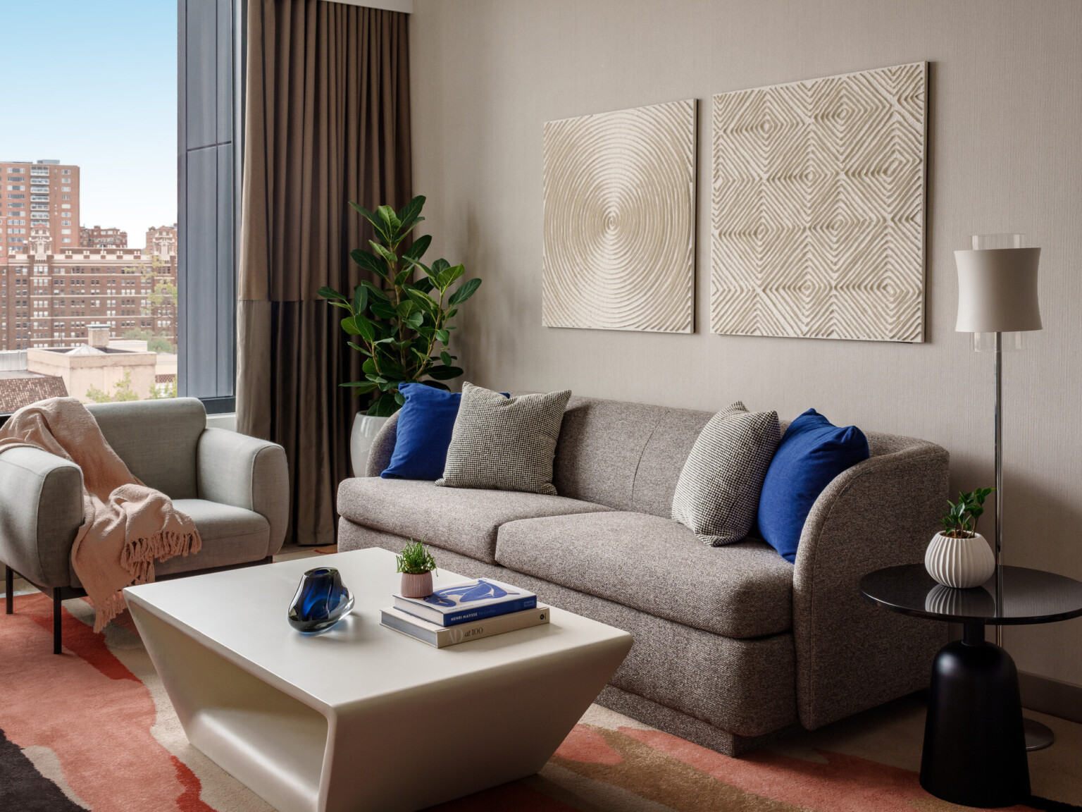 Modern hotel room sitting area showing neutral colored couch and chair with blue pillows, orange rug, and gold artwork hanging over the couch