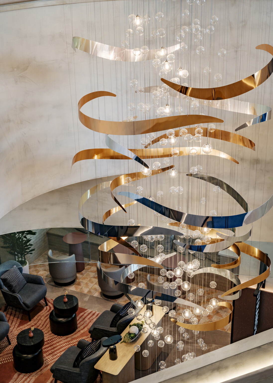 Stunning modern chandelier with cascading golden and silver ribbons, paired with clusters of glass globe lights, hanging above a stylish lounge filled with cozy chairs, round tables, and warm, textured rugs.