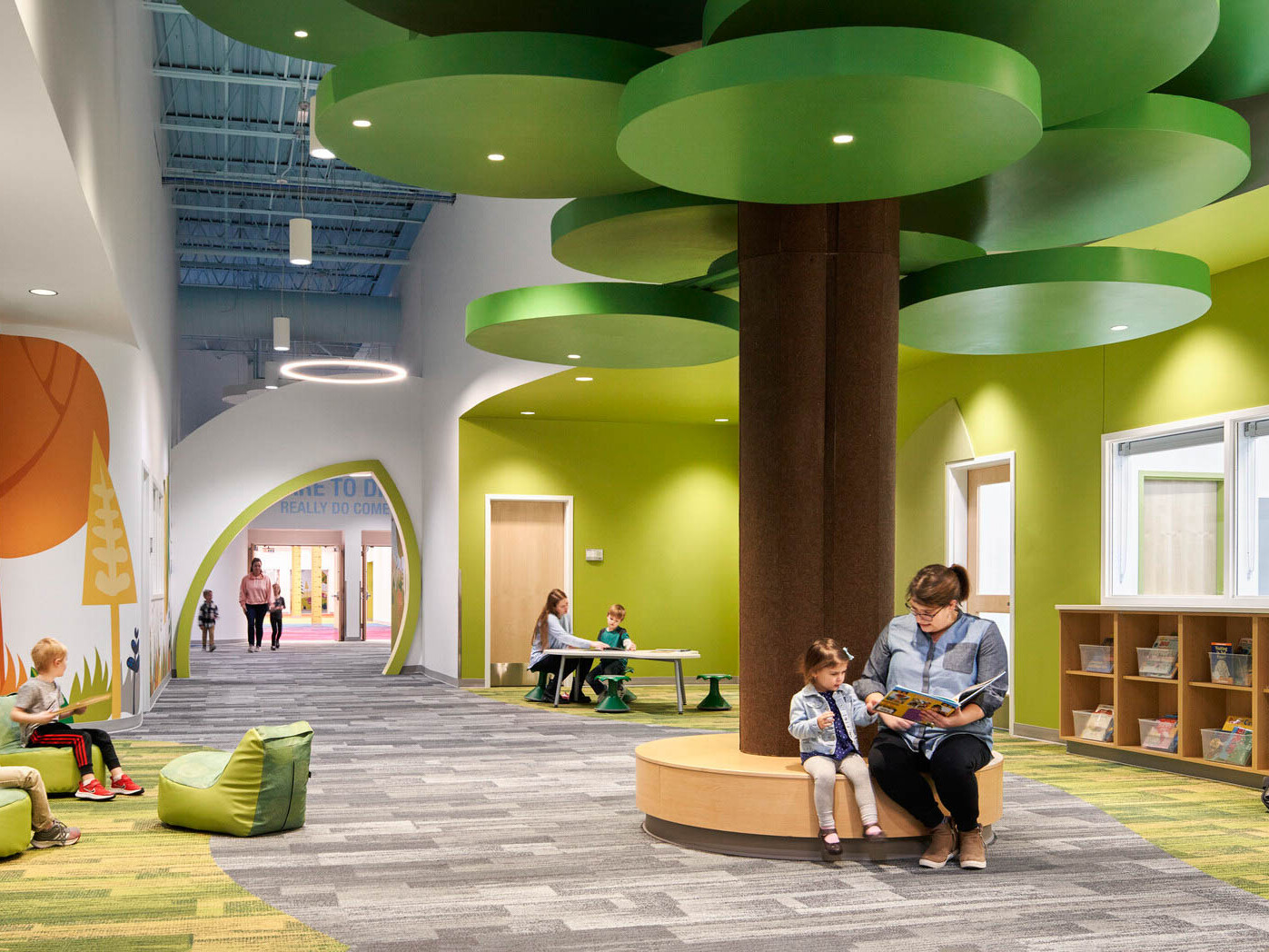 people reading on a brown circular bench with a brown column and green ceiling fixtures in hallway lined with forest graphics