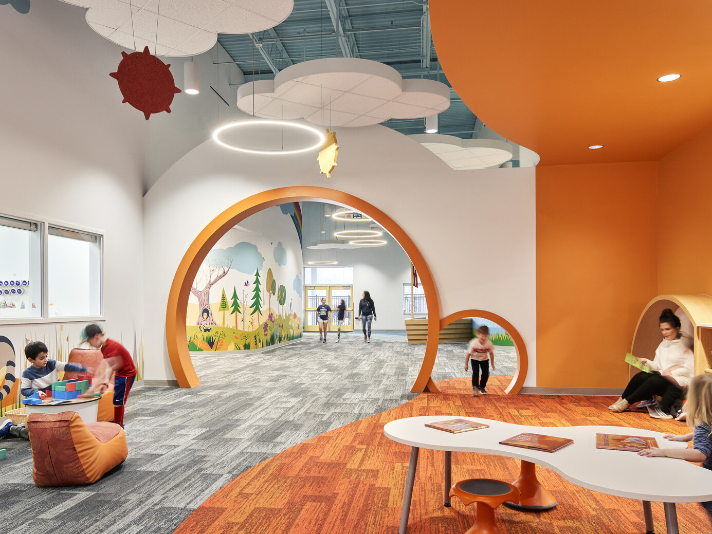 A vibrant children's play area with orange and grey décor, featuring playful murals, hanging decorations, and children engaging in activities.