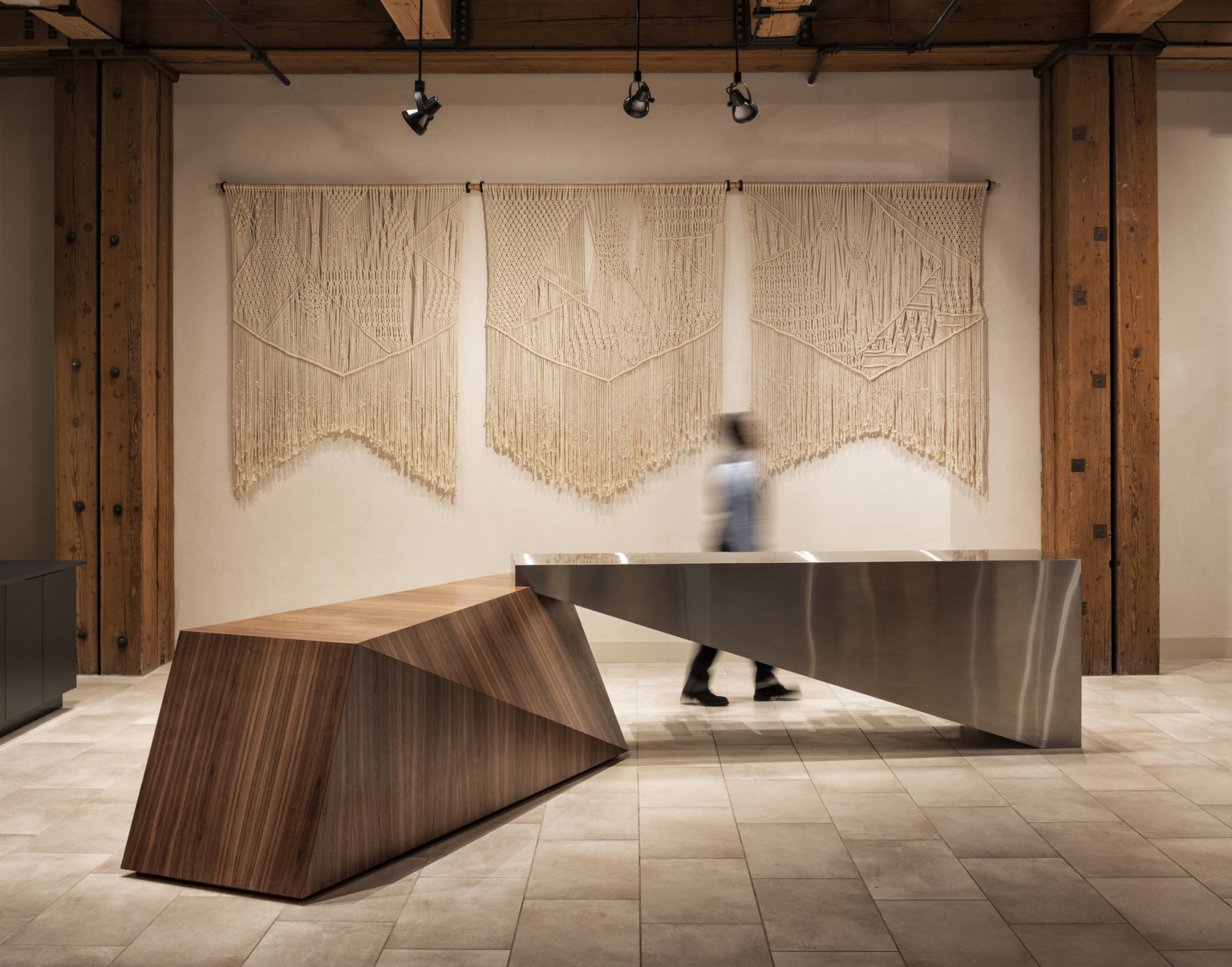 White wall with macrame wall hanging between wood pillars on tile floor with geometric wood and metal table at center