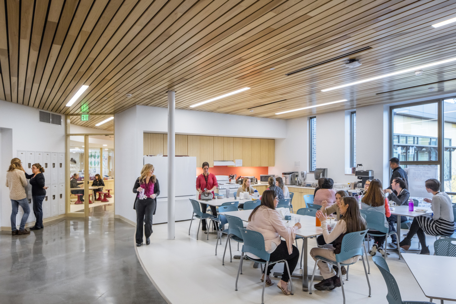 Workplace breakroom filled with tables and chairs with natural wood cabinets lining a back wall and floor to ceiling windows on the far wall