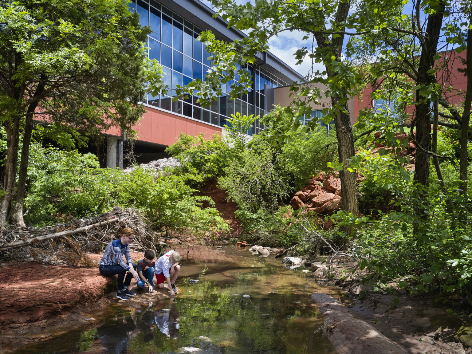 Two young students participating in outdoor learning near modern structure with floor-to-ceiling windows, lush landscaping and a reflecting pond