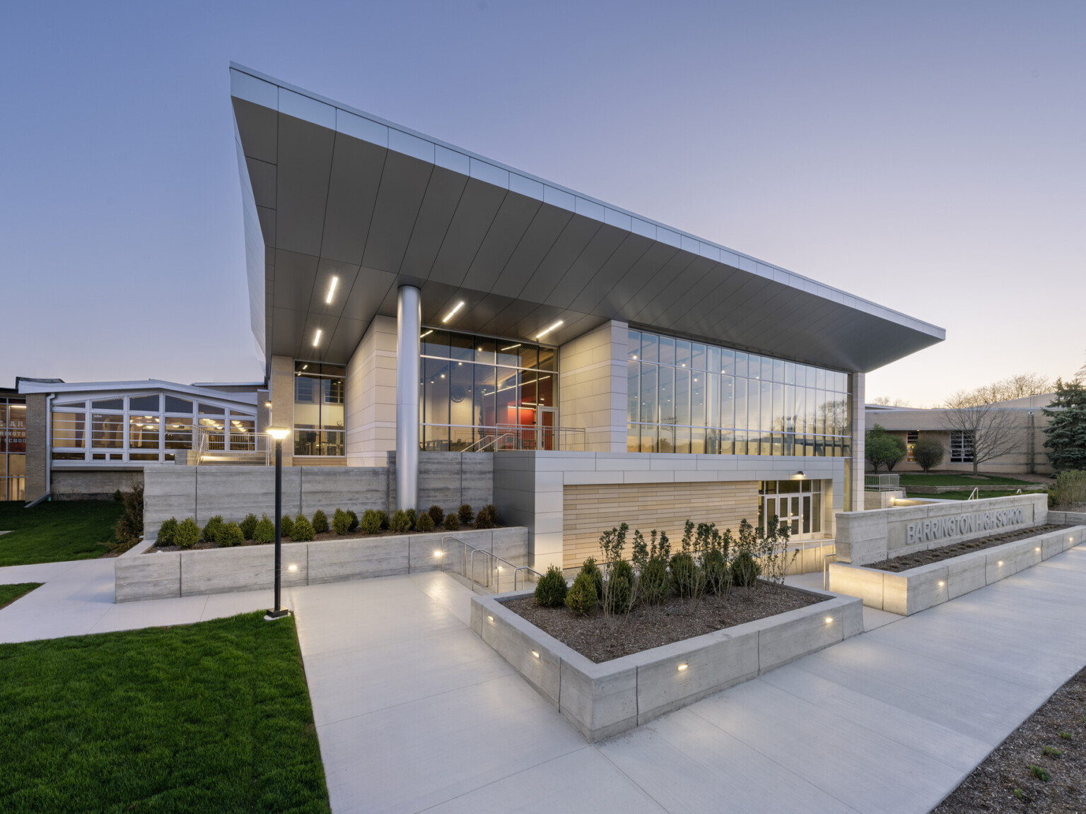 Exterior of Barrington High School showing a multistory building with lit up windows silver accents, and raised landscaping.