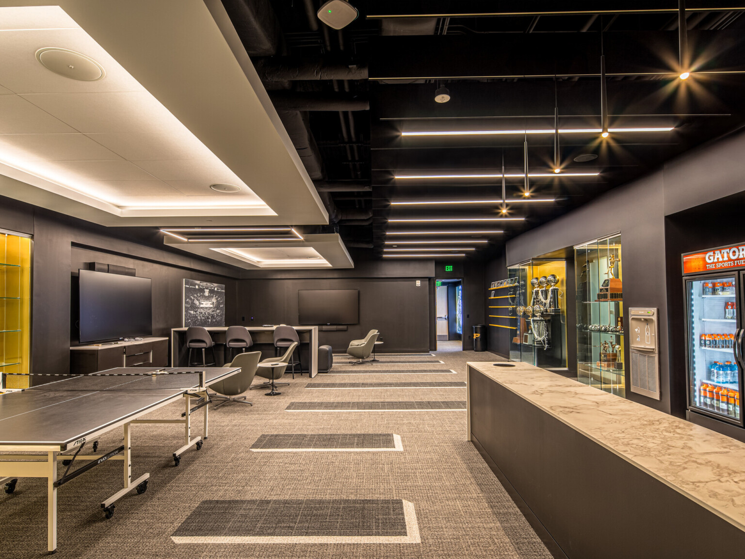 Recreation room with black walls, ping pong table, and multiple seating areas. Display cases along the walls filled with basketballs and trophies and Gatorade cooler