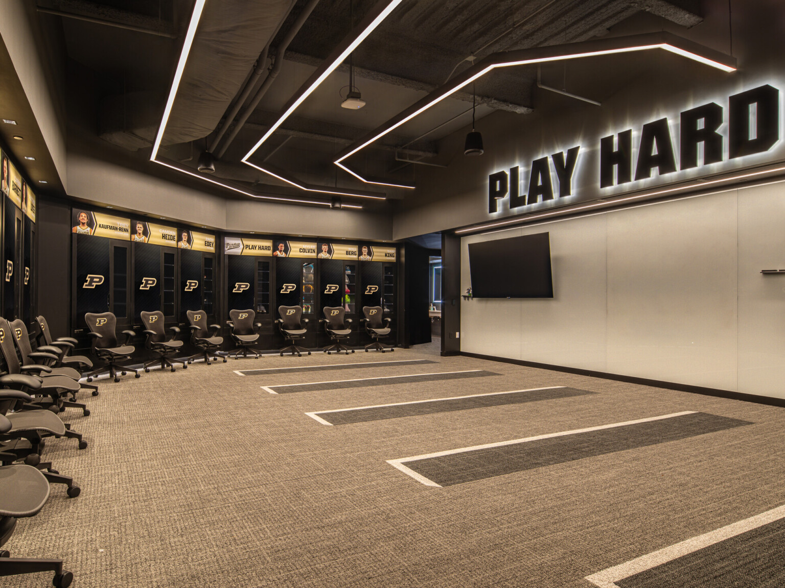 Purdue basketball locker room with wall lined with lockers and Purdue logo, TV hung on the right wall with words Play Hard backlit above