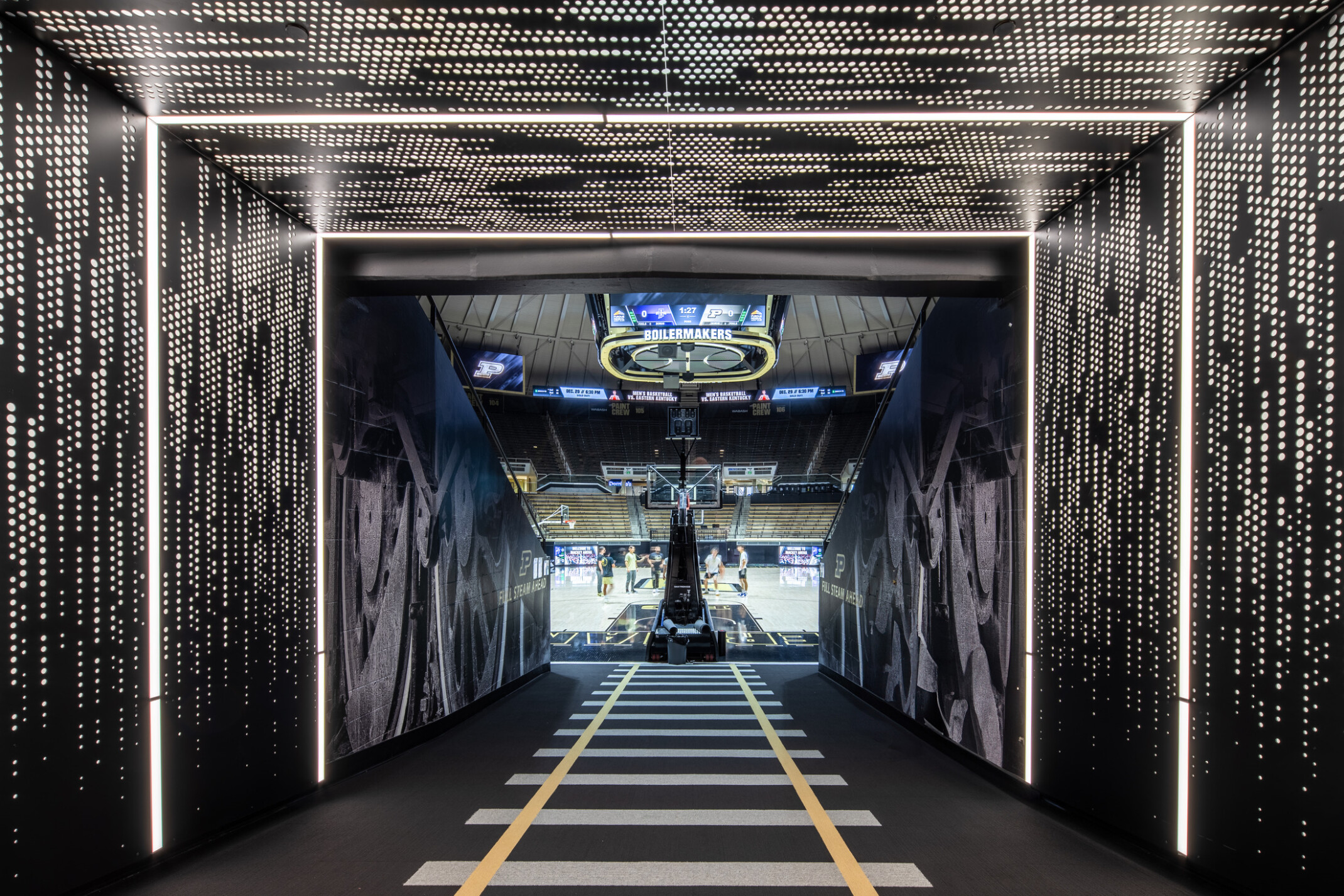 Tunnel at a basketball facility with patterned lights, sign from ceiling that says boilermakers, bench seating and basketball goal in the distance