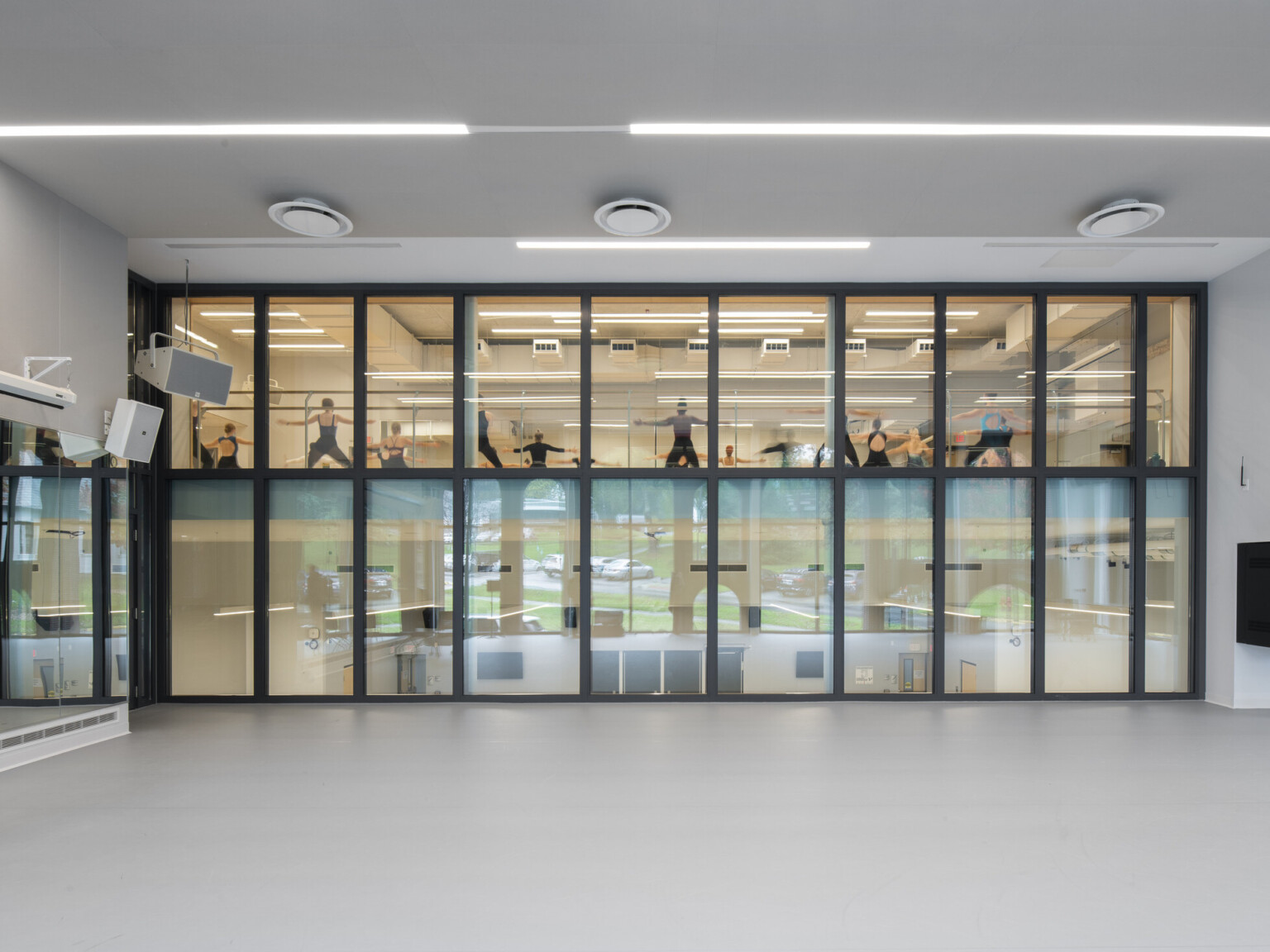 Large open room with grey floors and ceiling looking at large floor to ceiling windows outlined in black frames with a dance studio on the second floor filled with dancers.