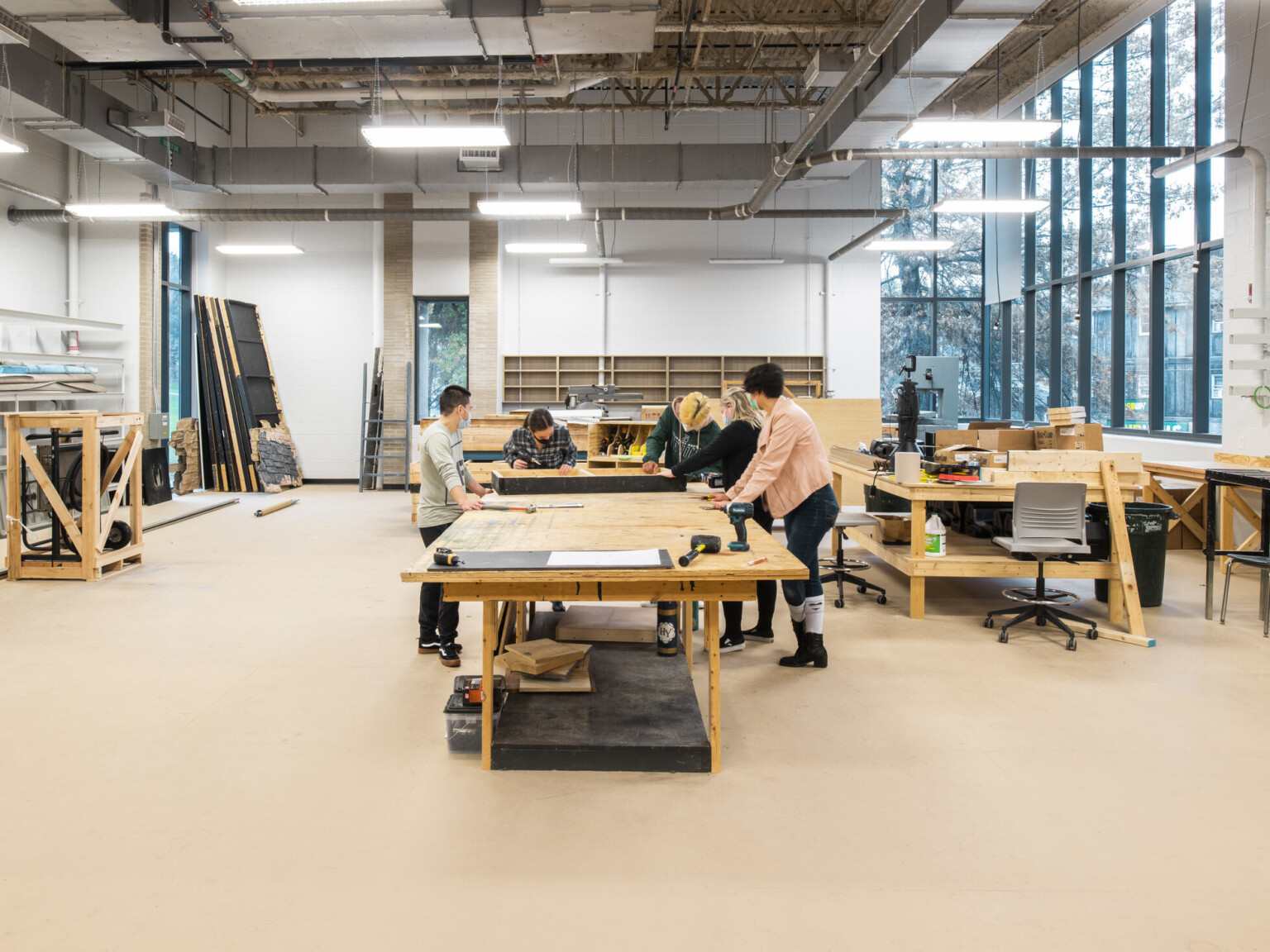 Construction classroom with wooden tables and building materials scattered throughout. A group of students observing someone building something in the center of the room