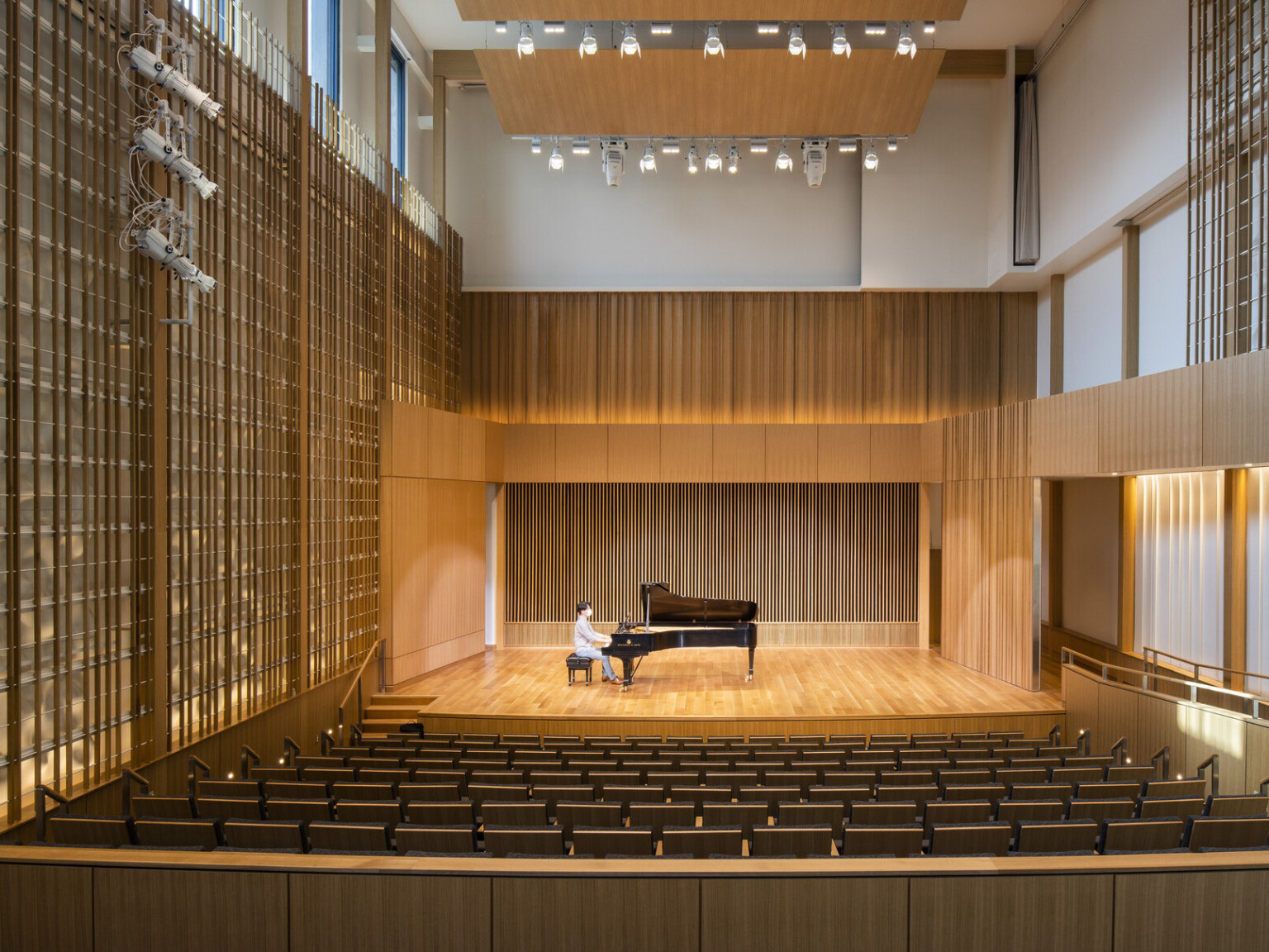 Man sitting at piano in double height recital room auditorium with theater seating. Wood panel designs on walls and wood acoustic panel