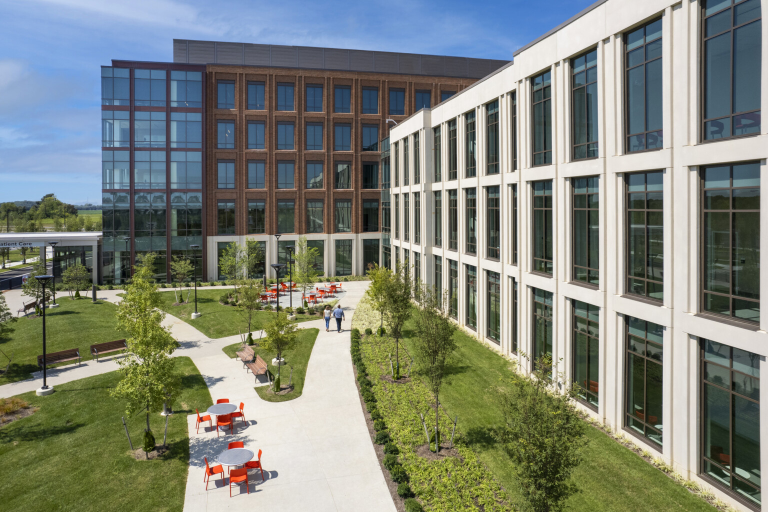 Aerial view of a concrete pathway along a white building surrounded by lush landscaping. Seating areas with grey table and orange chairs along with path