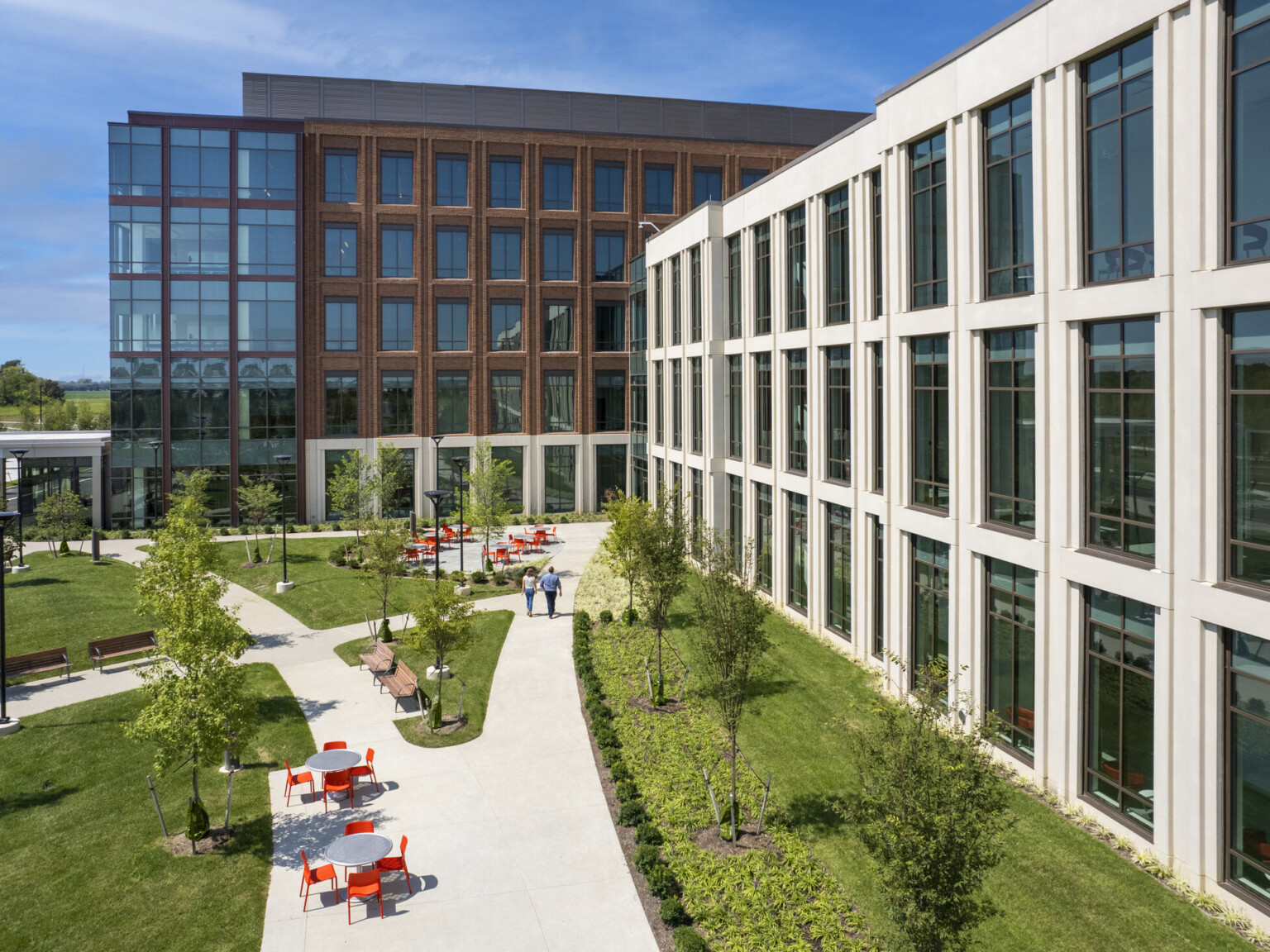 Aerial view of a concrete pathway along a white building surrounded by lush landscaping. Seating areas with grey table and orange chairs along with path