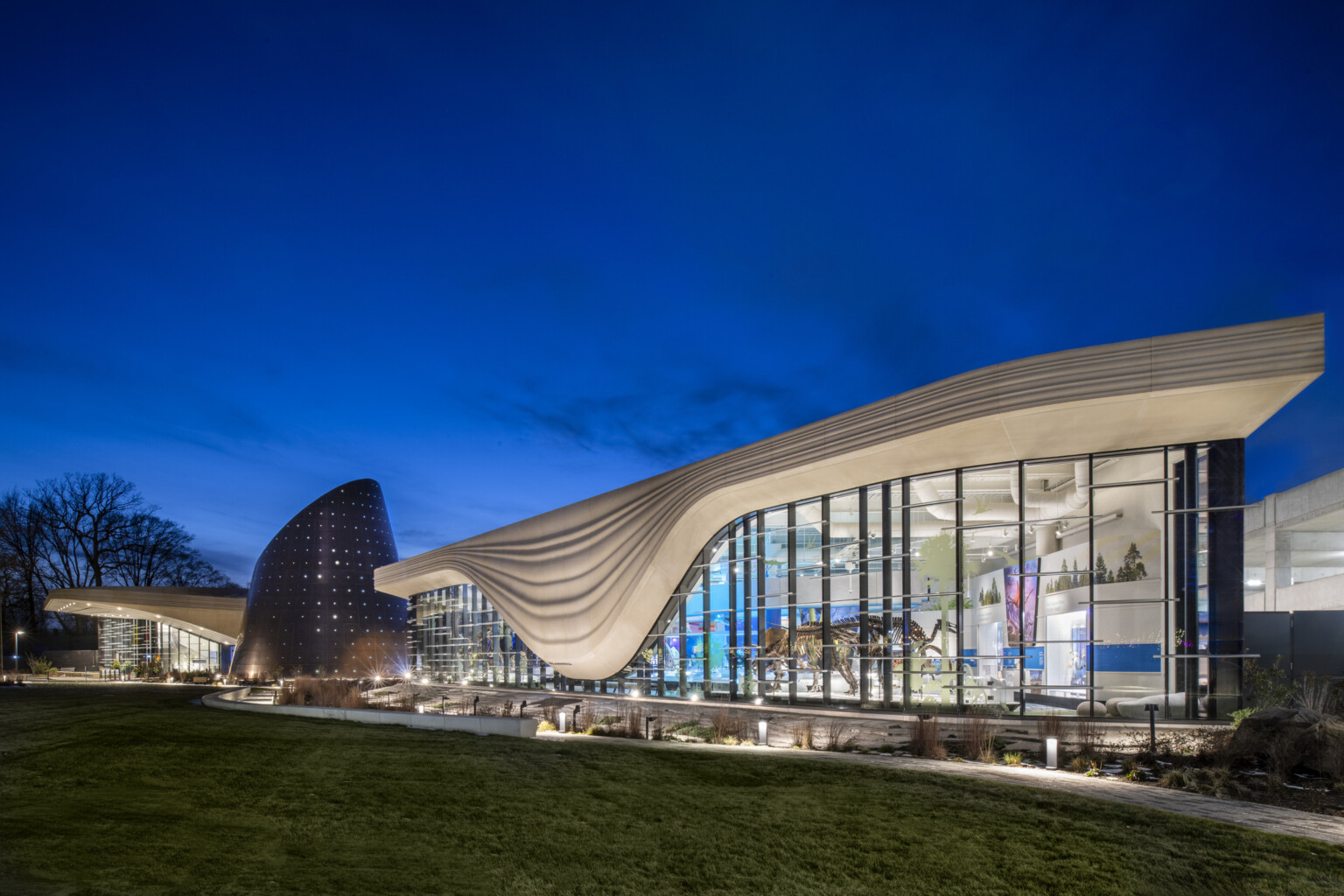 Nighttime image of a building with a white striped overhang that bows in center of right building section, an angled tube metal structure is at the building center