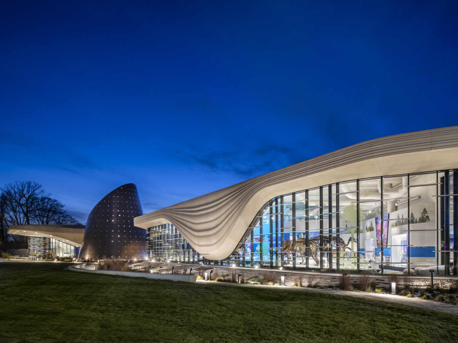 Nighttime image of a building with a white striped overhang that bows in center of right building section, an angled tube metal structure is at the building center