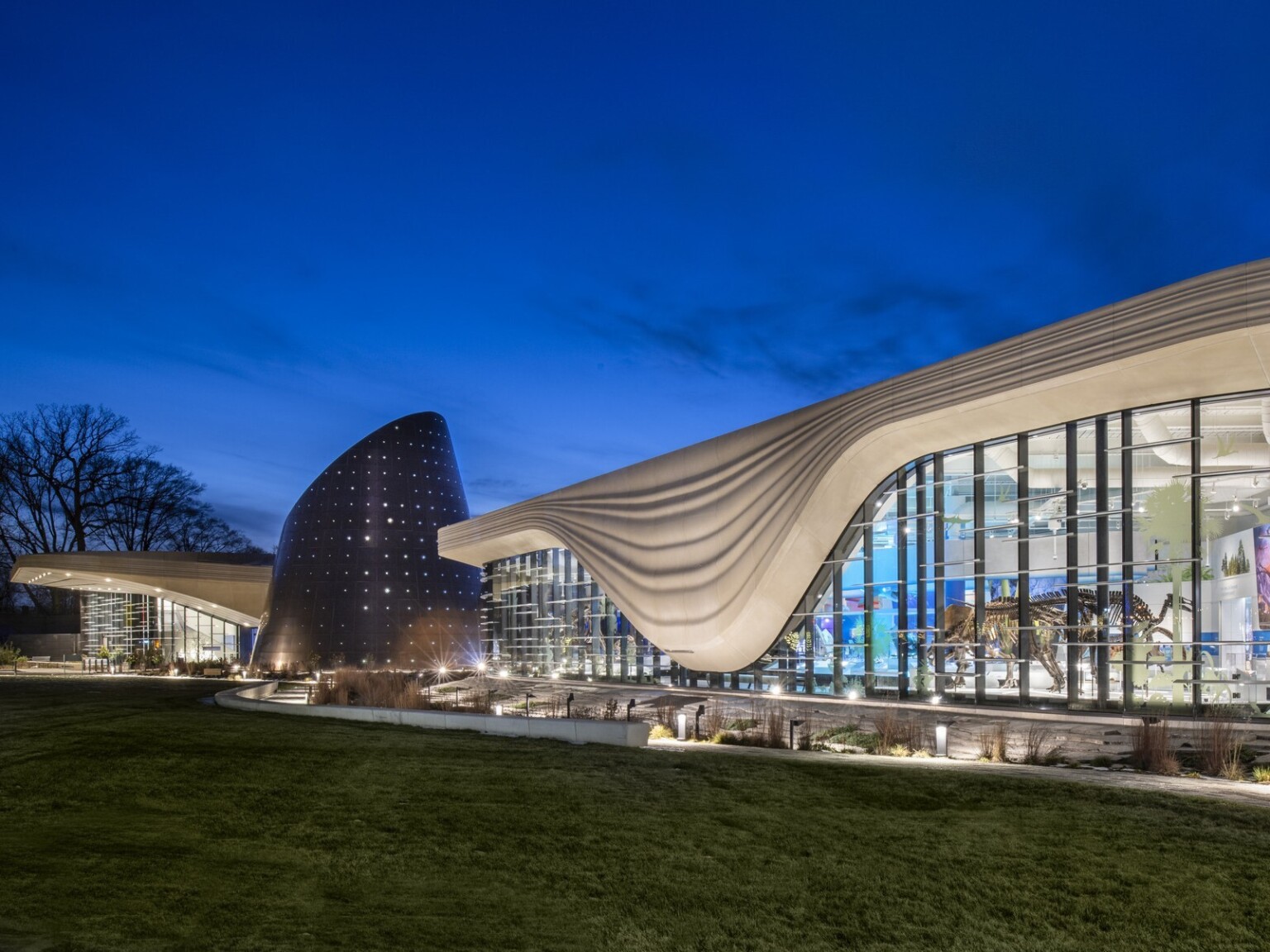 Nighttime image of a building with a white striped overhang that bows in center of right building section, an angled tube metal structure is at the building center