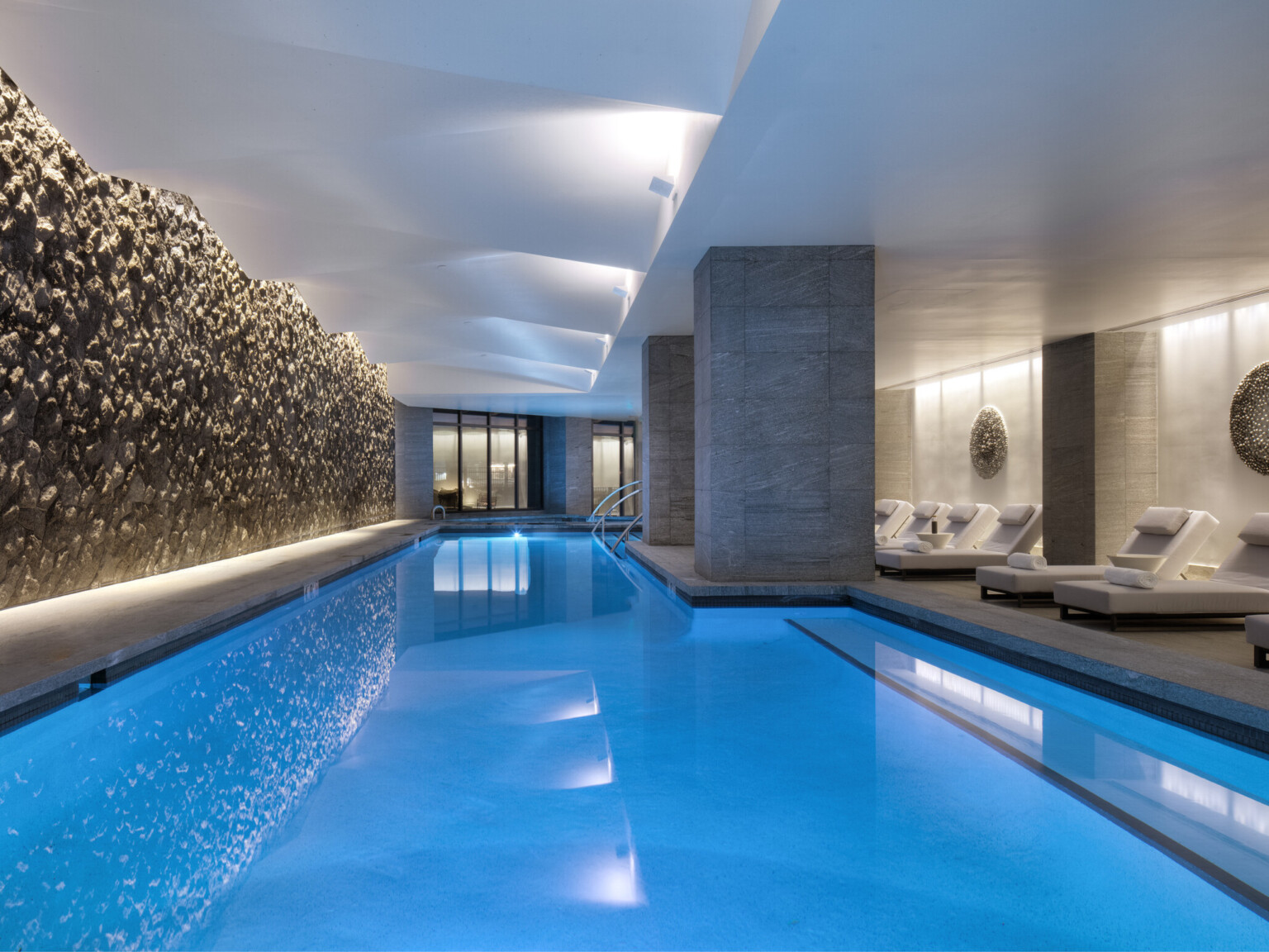 A hotel indoor pool lined with cushioned chaise loungers along a swimming pool lit up blue