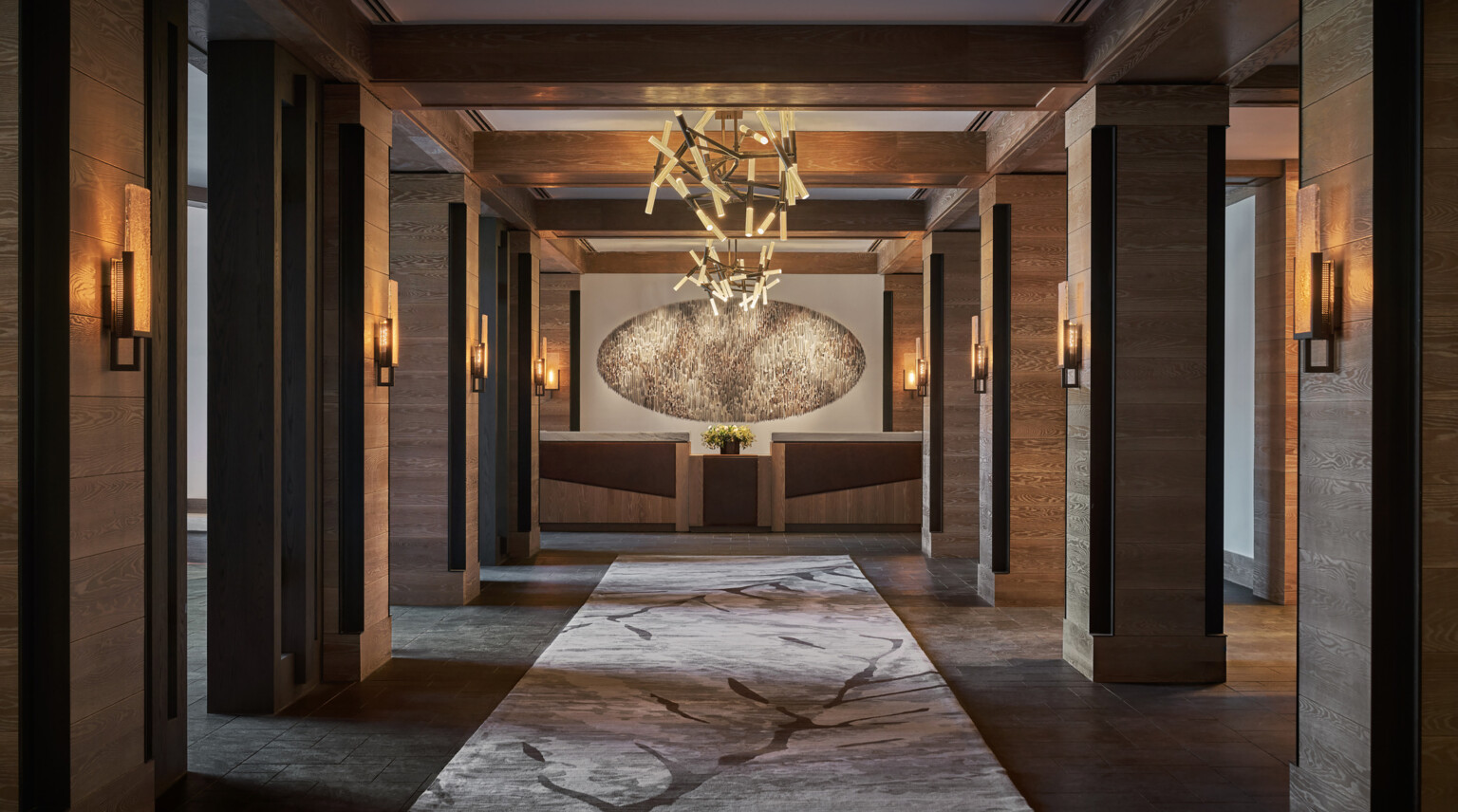 A modern hotel hallway with wooden columns with ambient lighting on each, and a central art piece at the end of the hallway