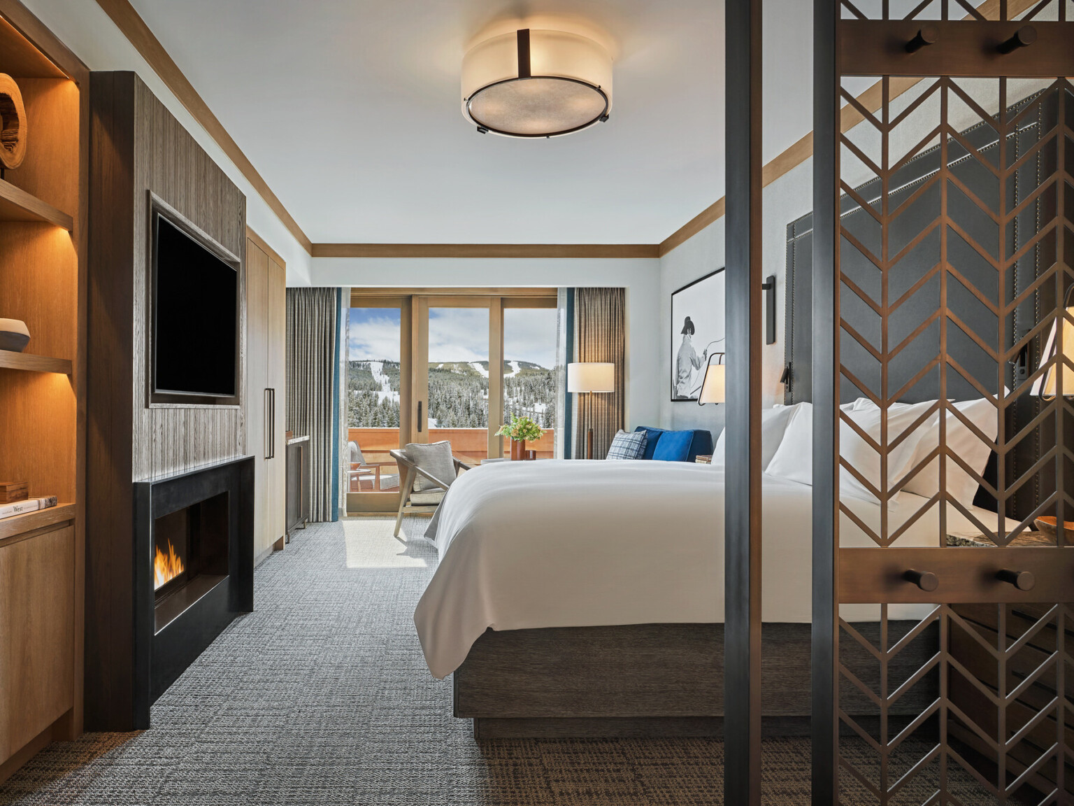 Modern hotel room with a large bed, balcony view of snowy mountains, and a fireplace under a mounted TV