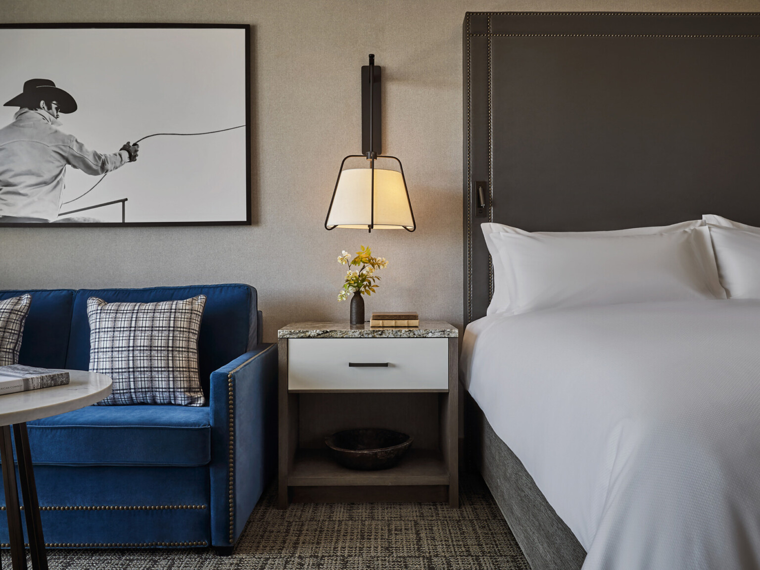 Hotel room with a blue sofa with nail head details, large cowboy photo, bedside table with flowers, and a neatly made bed with crisp white linens