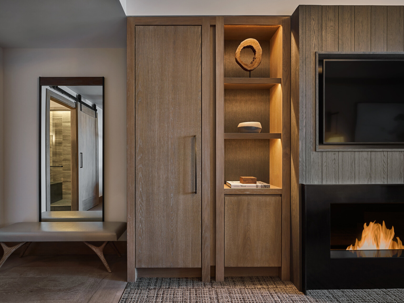 Modern hotel room with wooden cabinets, shelves with lighting, a large rectangular mirror with bench underneath, and a TV mounted over a fireplace