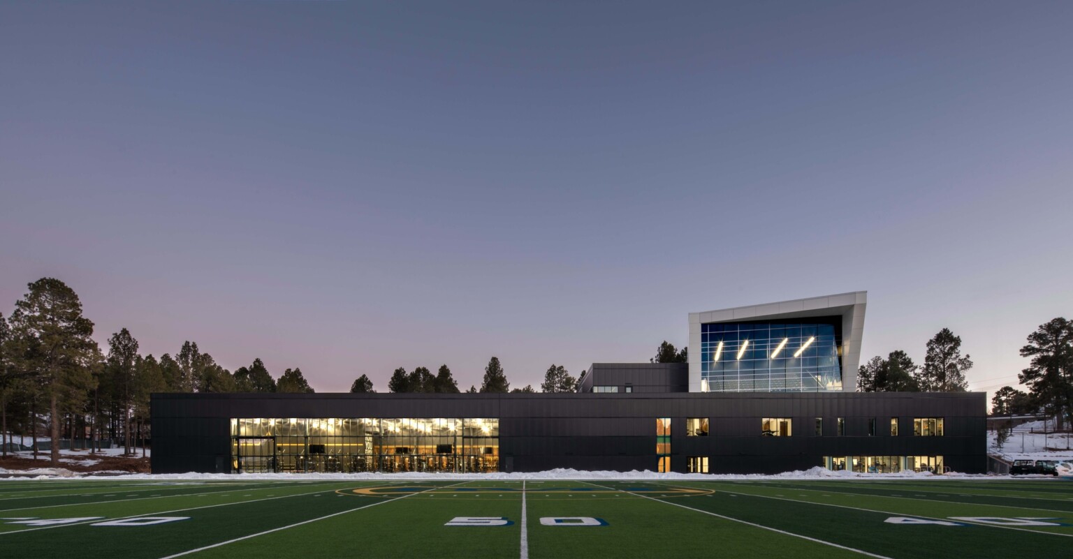 football field in front of a single story building with a multi story angular volume behind it against tree line sky at dusk at Northern Arizona University