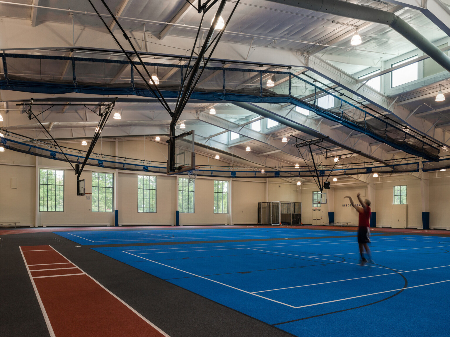 Indoor basketball court with a blue floor surrounded by black and red accents with exposed beams and windows in the ceiling