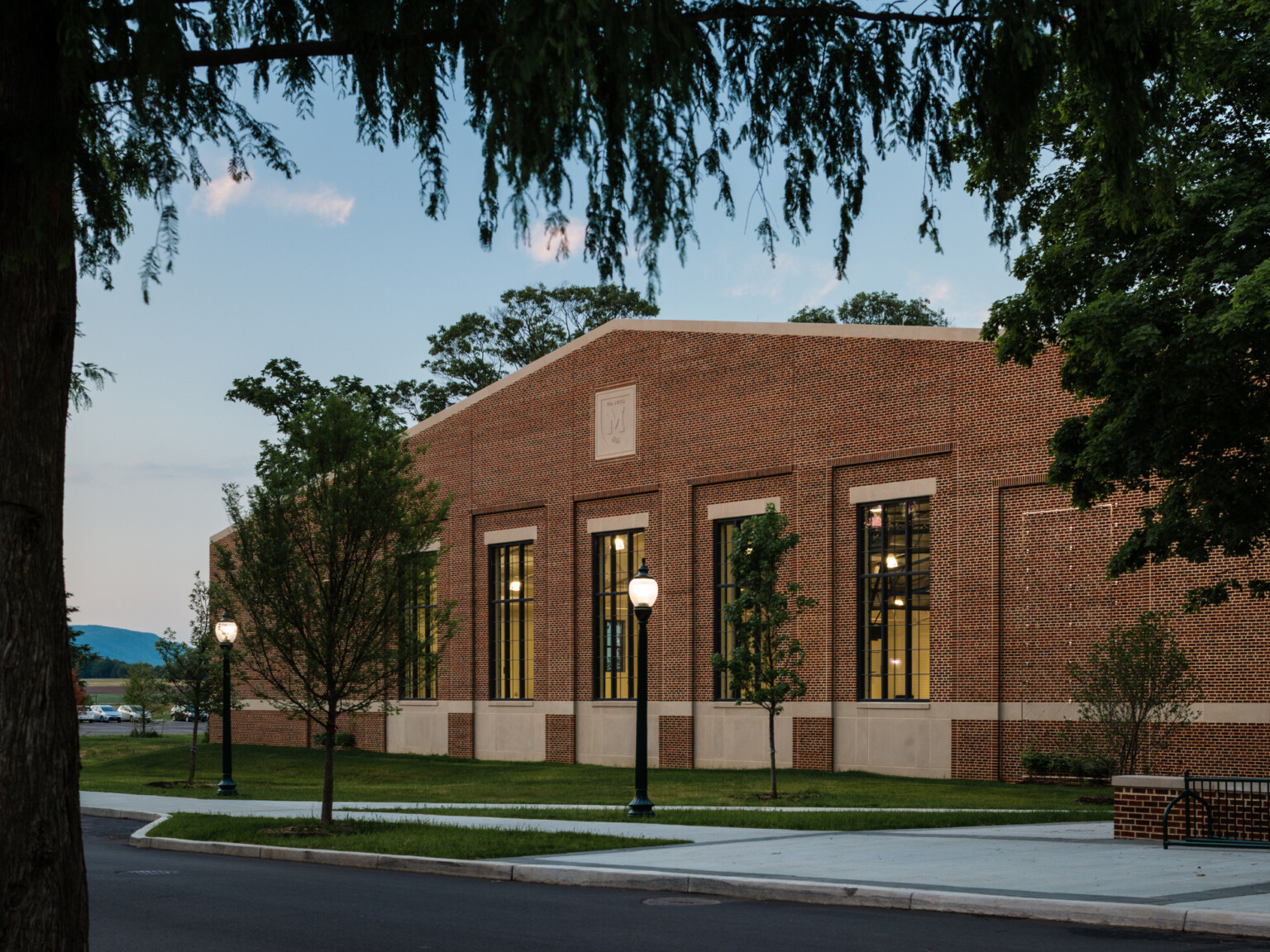 Brick building with multiple large windows, sidewalk lined with lighting and green trees