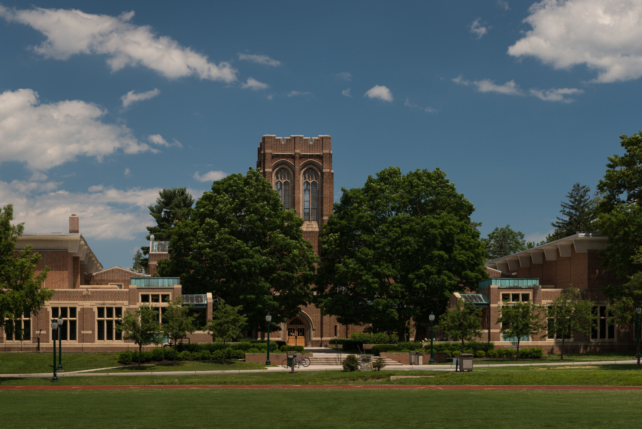 Exterior of s brick school with a large center tower filled with ornate windows flanked by large green trees and smaller side buildings with green roofs