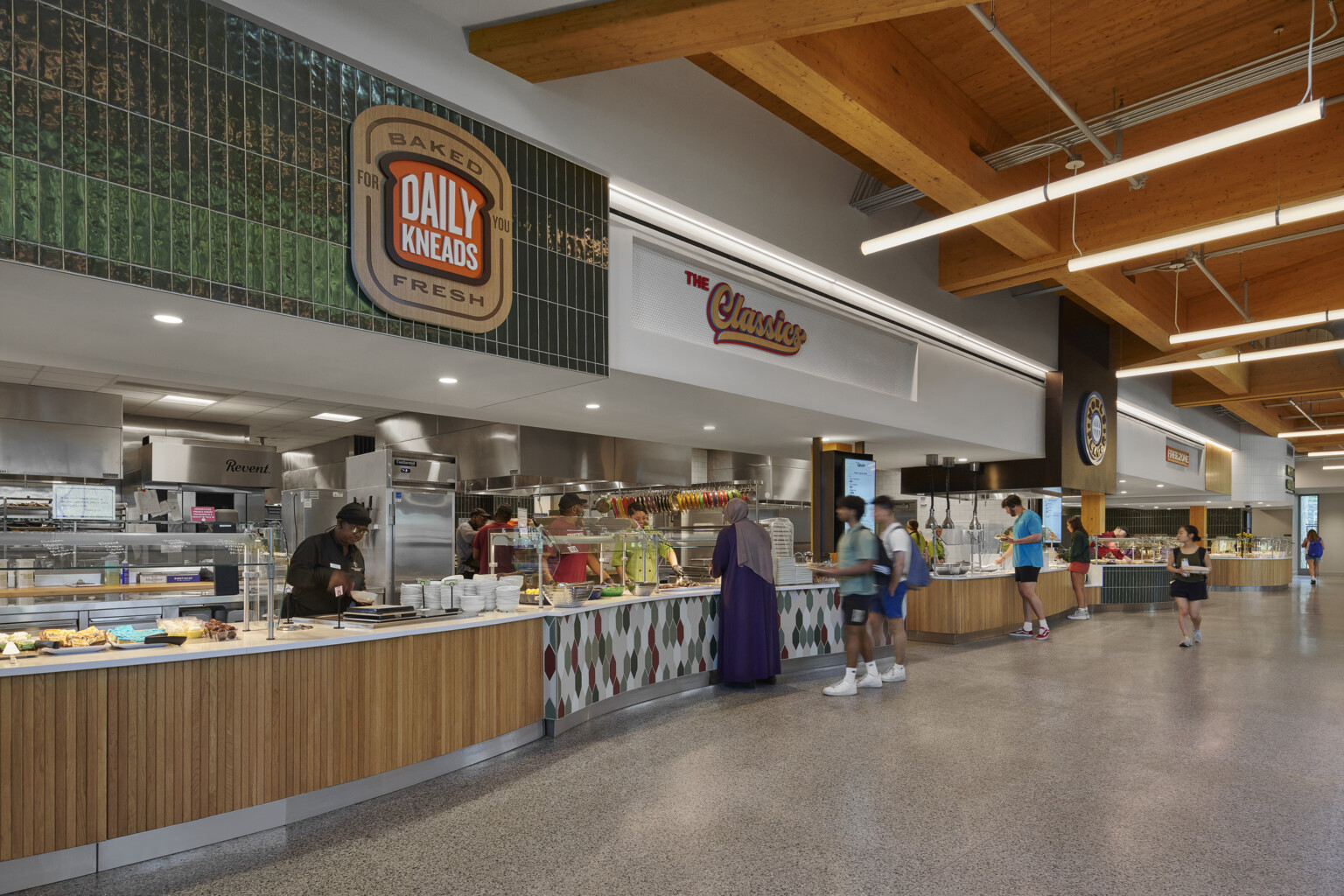 College dining hall with tall ceilings, exposed mass timber wood ceiling, green and white tile accents, raw concrete floors