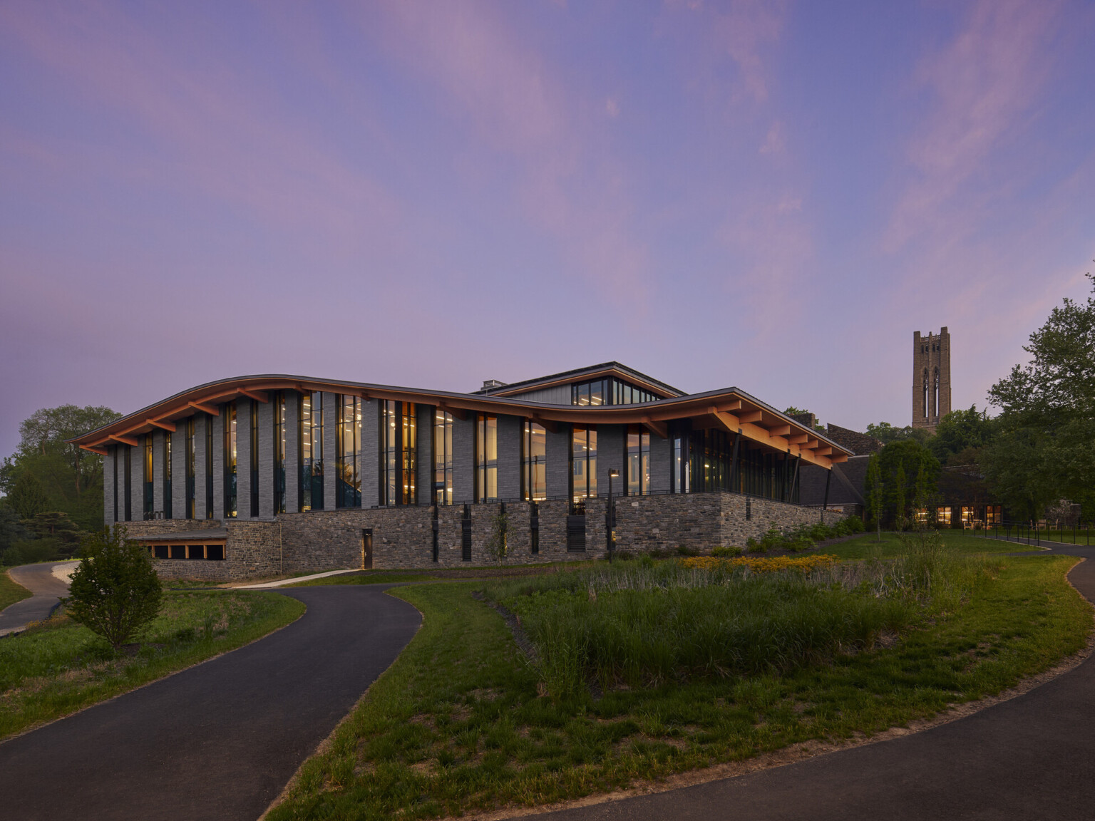 Stone and glass facade on university building with curving roof and overhang illuminated from within in the evening, mass timber architecture, what is mass timber construction