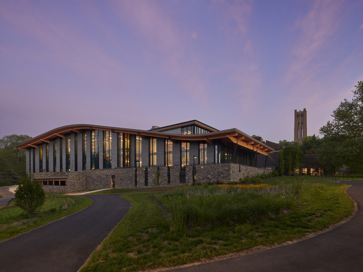 Stone and glass facade on university building with curving roof and overhang illuminated from within in the evening, mass timber architecture, what is mass timber construction