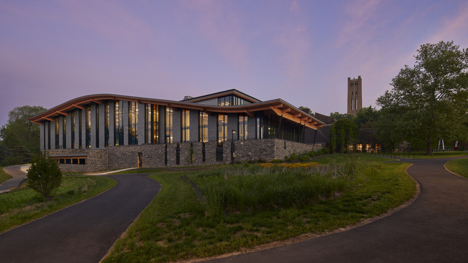 Stone and glass facade on university building with curving roof and overhang illuminated from within in the evening, mass timber architecture, what is mass timber construction