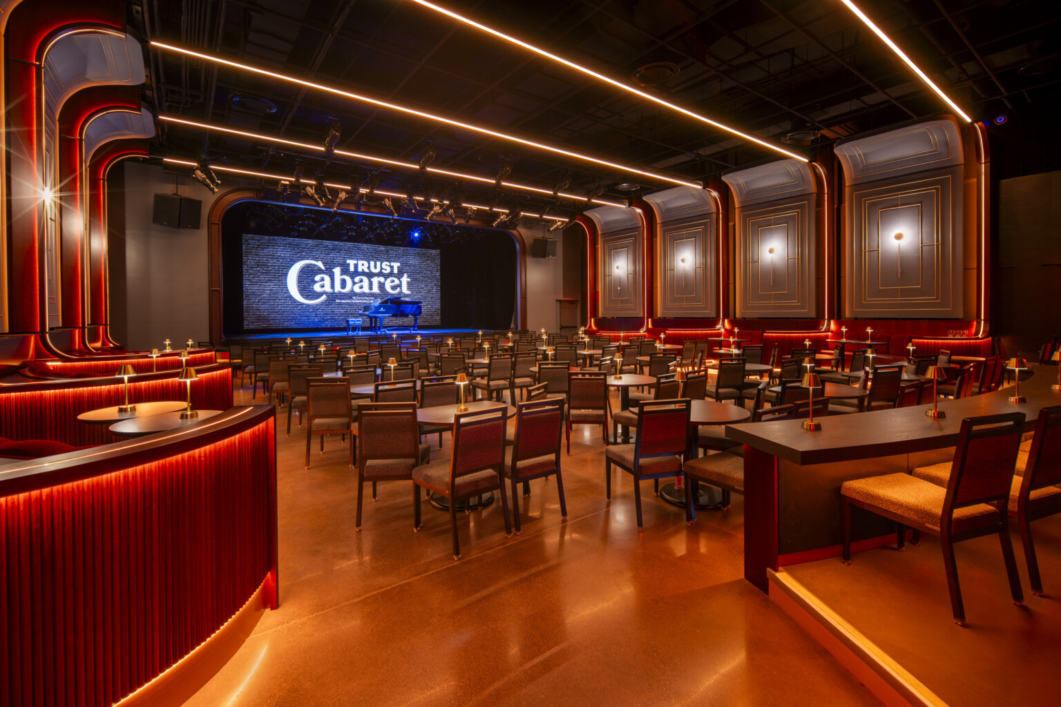 Lounge filled with tables and chairs facing a stage with a piano in the middle. Ceiling and walls accented by neon lights.