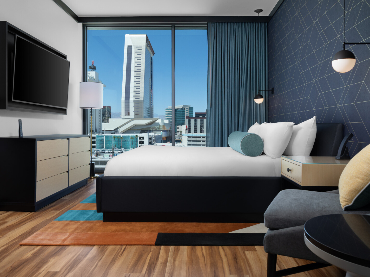 Modern hotel room with wooden floors, floor to ceiling window overlooking high rise buildings, dark blue accent wall