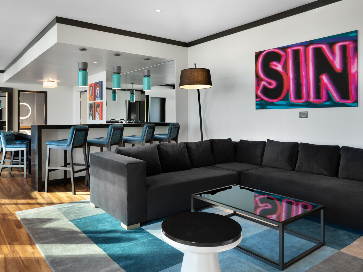 Living area with light wood floors, dark couch, bar with blue upholstered bar stools, paining on wall with word SIN