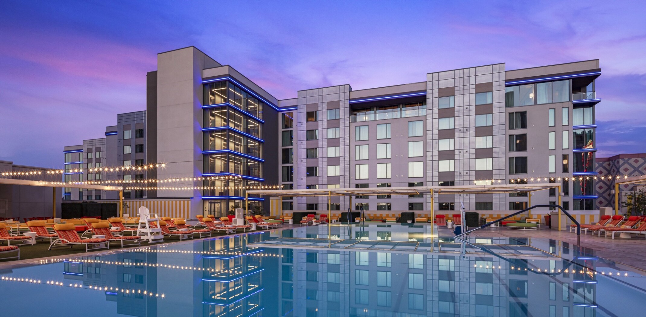 Modern multistory building with blue accent lighting outlining glass windows behind a large pool surrounded by orange tanning chairs