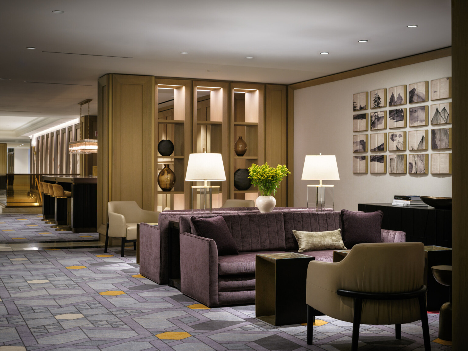 Hotel lobby seating area filled with luxurious purple couches, light wood shelves filled with vases, bar area with gold bar stools