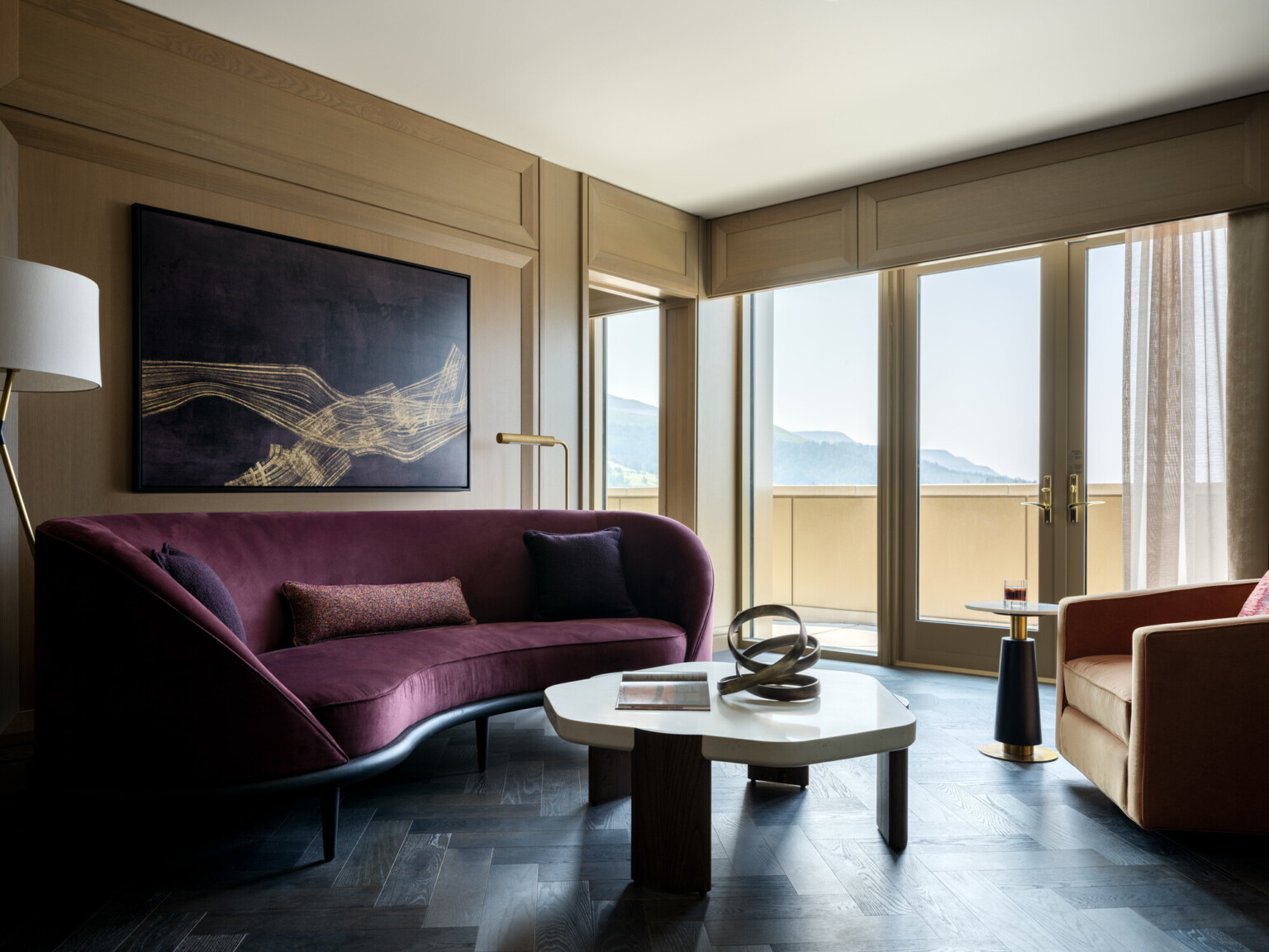 Curved high back purple couch, floor to ceiling windows with doors, black herringbone flooring, gold curtains