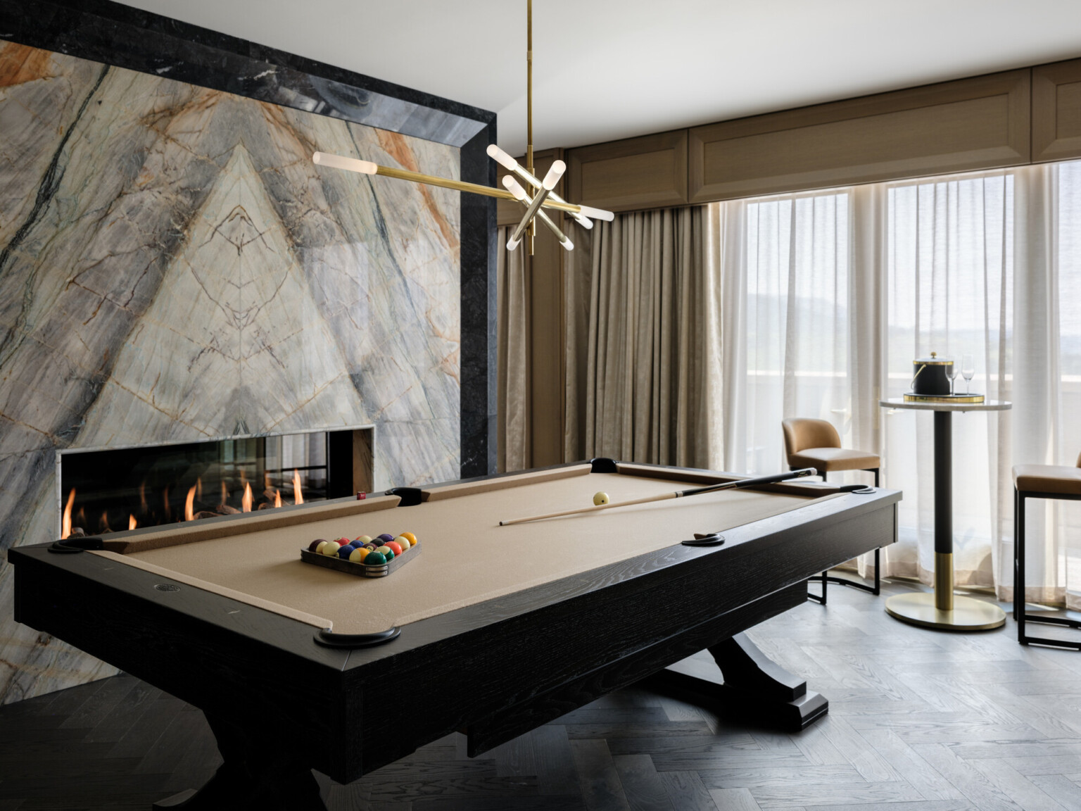 Modern pool table with modern gold light fixture above in front of a marble fireplace, high table and bar stools