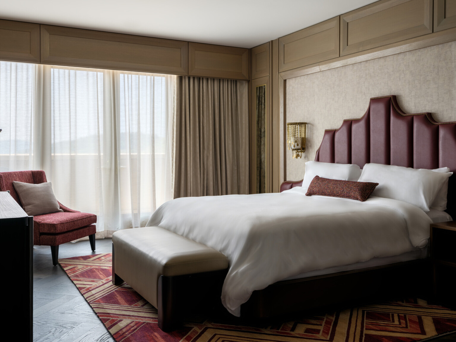 Hotel room with bed with red headboard and soft white linens, red accent chair, large windows covered by sheer curtains