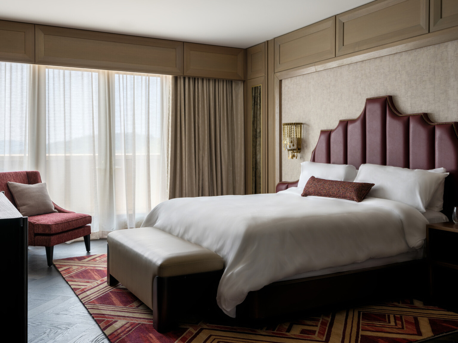 Hotel room with bed with red headboard and soft white linens, red accent chair, large windows covered by sheer curtains