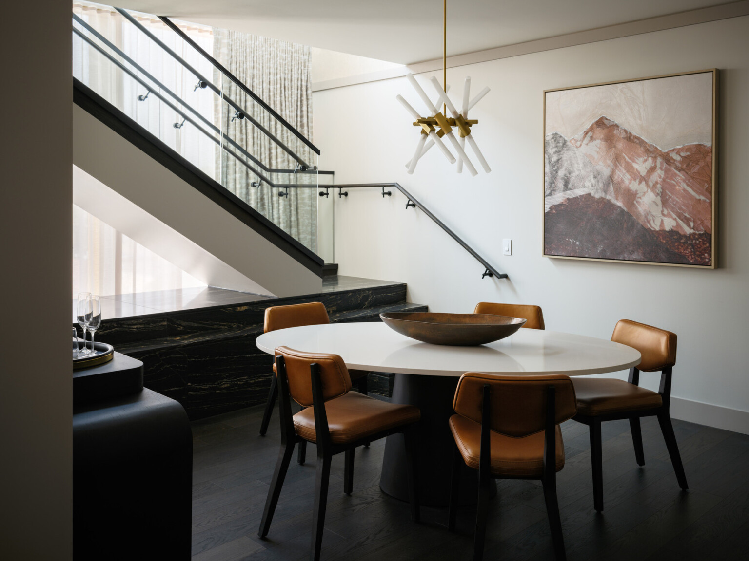 Seating area with white table and copper colored chairs, modern staircase with glass railings, modern painting in shades of orange